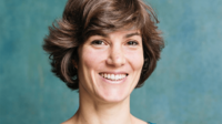 Elena Hassinger mit kurzen braunen Haaren und hellem Lächeln vor blauem Hintergrund.